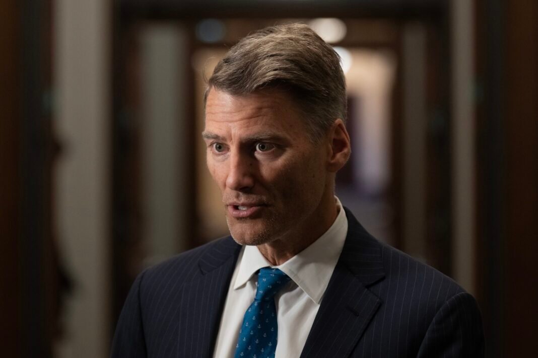 A close-up of Gregor Robertson wearing a dark suit with a blue tie. He is speaking to someone off to the camera's left.
