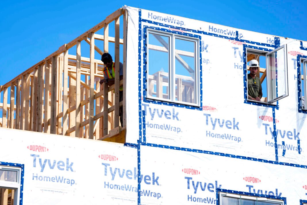 Workers work on a house frame wrapped in plastic against a clear blue sky.